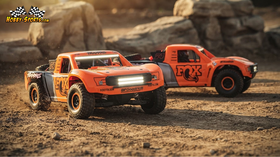 Two orange and black Traxxas Unlimited Desert Racer RC trucks kicking up dust racing side by side on a rocky desert trail.