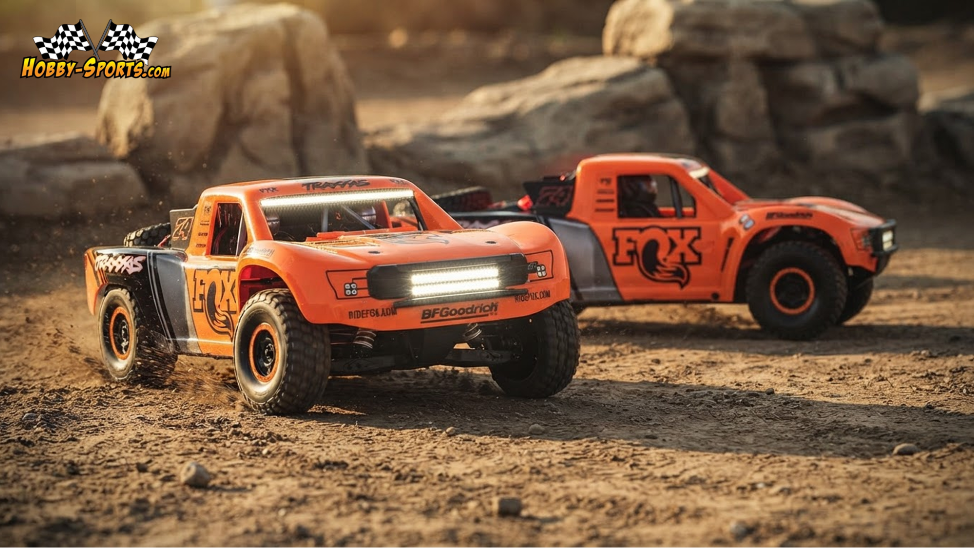Two orange and black Traxxas Unlimited Desert Racer RC trucks kicking up dust racing side by side on a rocky desert trail.
